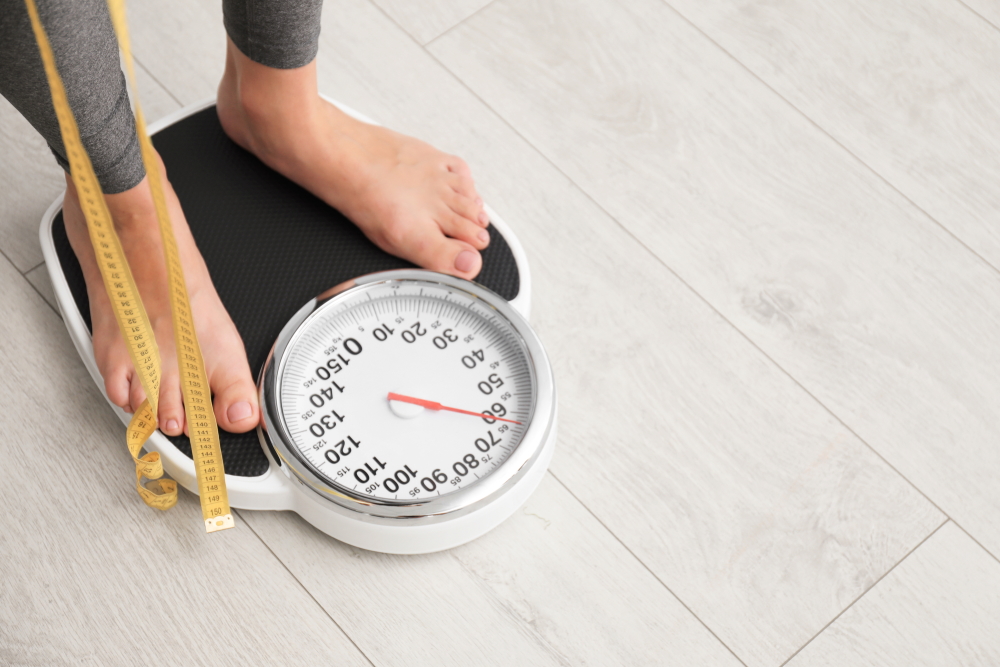 Žena stojí na váze s metrem v ruce a měří si hmotnost