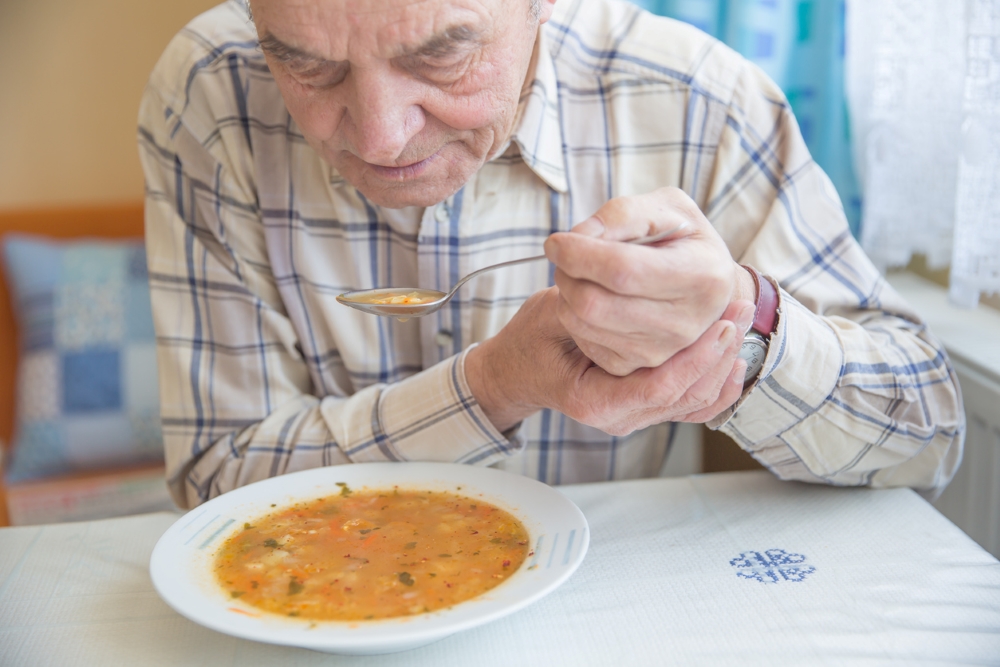 Starý muž jí polévku a ruku se lžící si přidržuje druhou