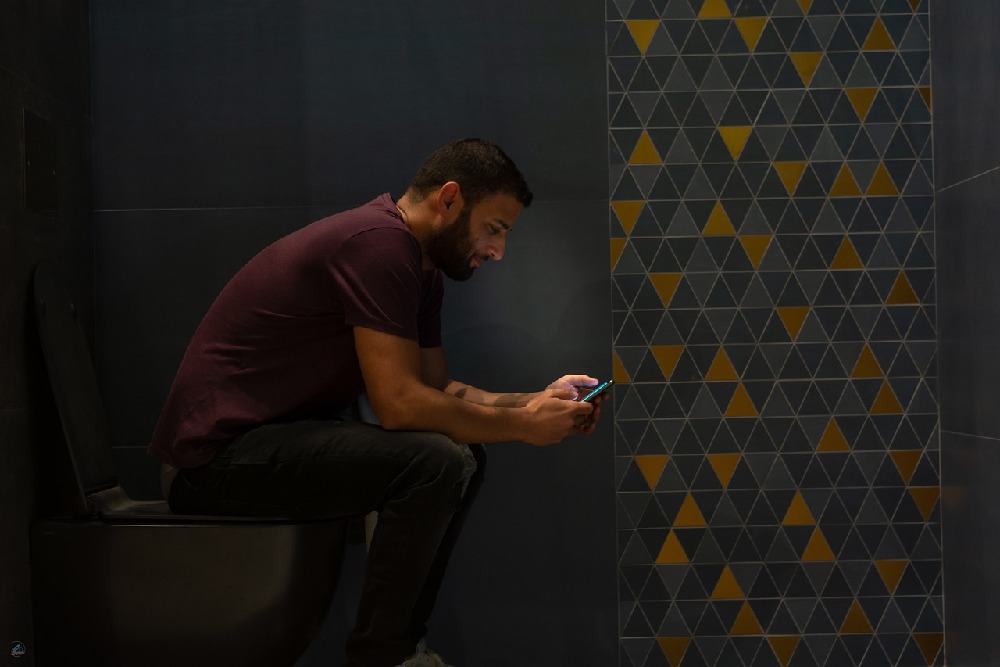 Muž sedící na záchodě v šatech kouká do mobilního telefonu