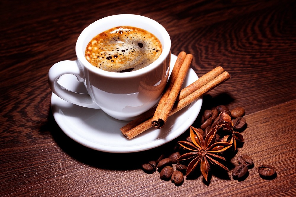 Káva s přidanou skořicí v bílém hrnku na bílém podšálku dekorativně ozdobená tyčinkami skořice, kávovými zrny a badyánem