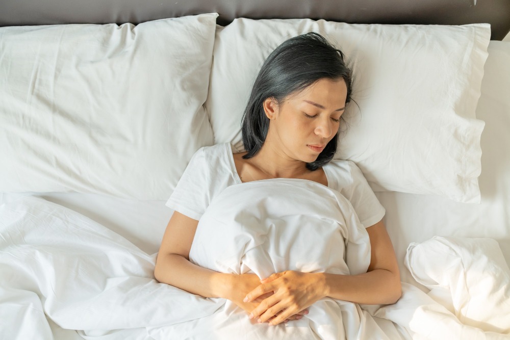 Mladá žena trpící chronickou únavou při onemocnění ledvin ležící v posteli s rukama přes podbřišek.