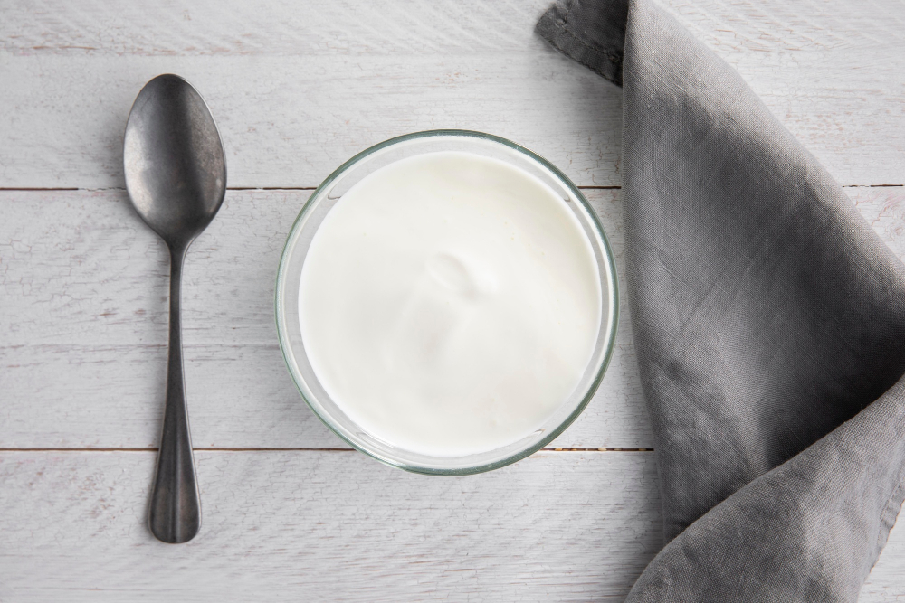 Bílý jogurt ve skleněné mističce s připravenou lžící a šedým látkovým ubrouskem na dřevěném stole
