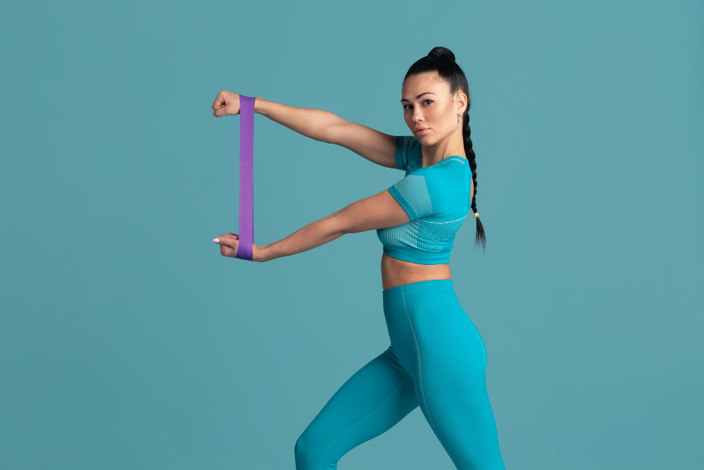 Žena v modrém cvičebním oděvu s fialovou gumou mezi rukama, provádějící cvik k posílení zad a podpaží