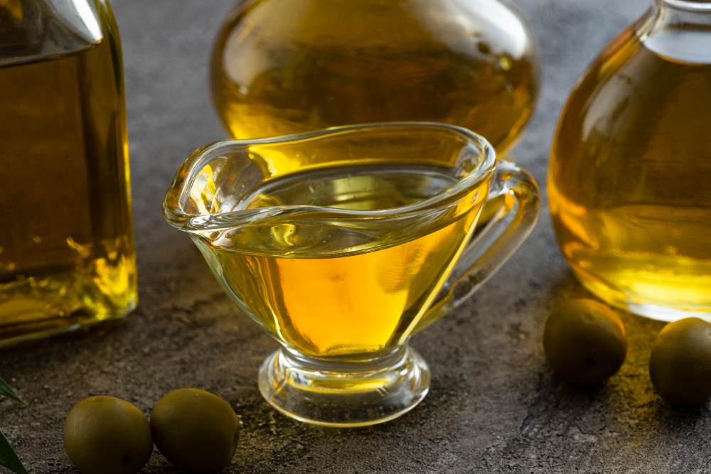 Skleněný hrneček s olivovým olejem, v pozadí skleněné karafy s olivovým olejem a olivy na stole kolem hrnku