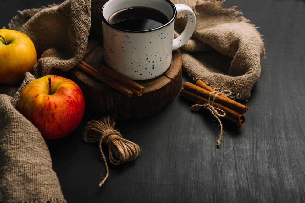 Čaj se skořicí v plechovém hrnku na podložce ze dřeva na stole s provázkem, skořicí a dvěma jablky