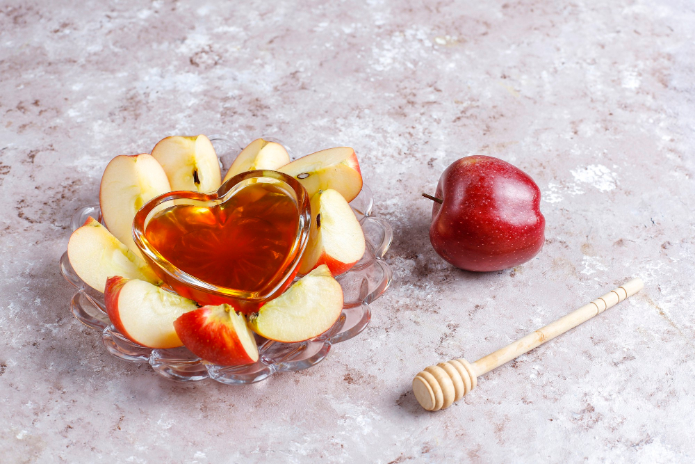 Mistička s medem ve tvaru srdce obklopená nakrájeným jablkem, vedle celé jablko a dřevěné nabírátko na med