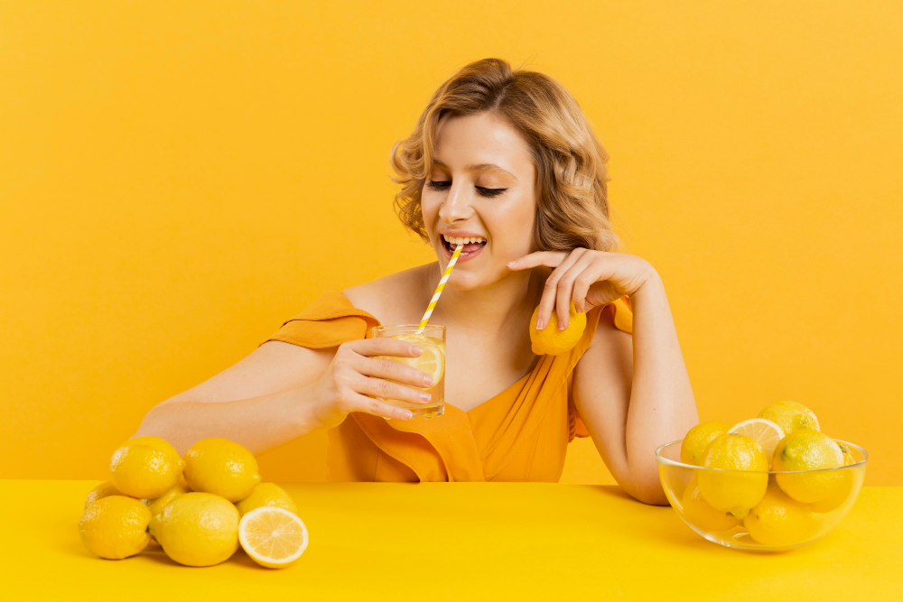 Žena popíjející brčkem citronovou vodu ze sklenice, v ruce drží citron a před sebou má misku plnou citronů a hromadu citronů na stole