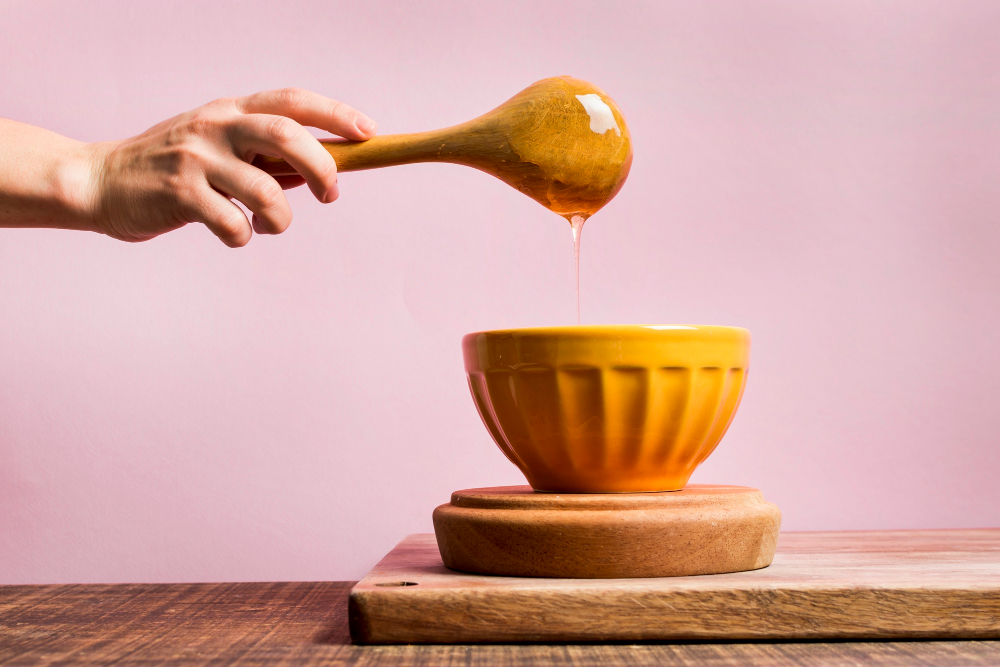 Porcelánová žlutá miska na prkénku, do které stéká med z dřevěné vařečky, kterou drží ženská ruka ve vzduchu