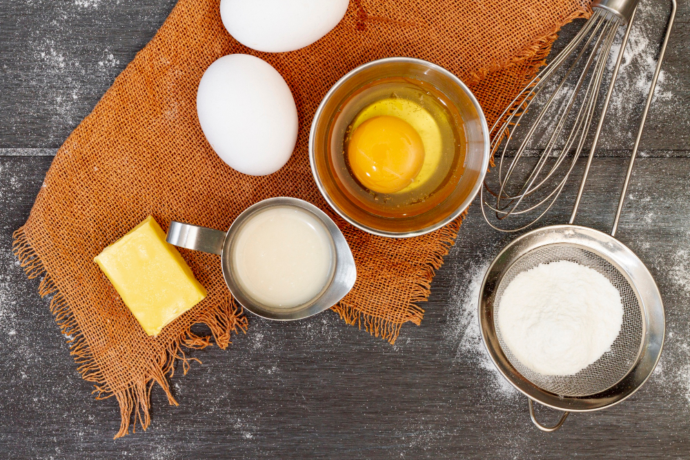 Kus másla, mléko v odměrce, žloutek ve sklenici a dvě bílá vejce na stole s metličkou