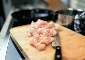 Nakrájené syrové kuřecí maso na kostičky na dřevěném prkénku s nožem v kuchyni