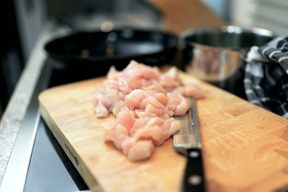 Nakrájené syrové kuřecí maso na kostičky na dřevěném prkénku s nožem v kuchyni