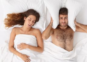 Muž ucpávající si polštářem uši, vedle spící a chrápající partnerka
