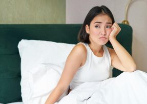 Mladá žena sedící v posteli se zkroušeným výrazem ve tváři