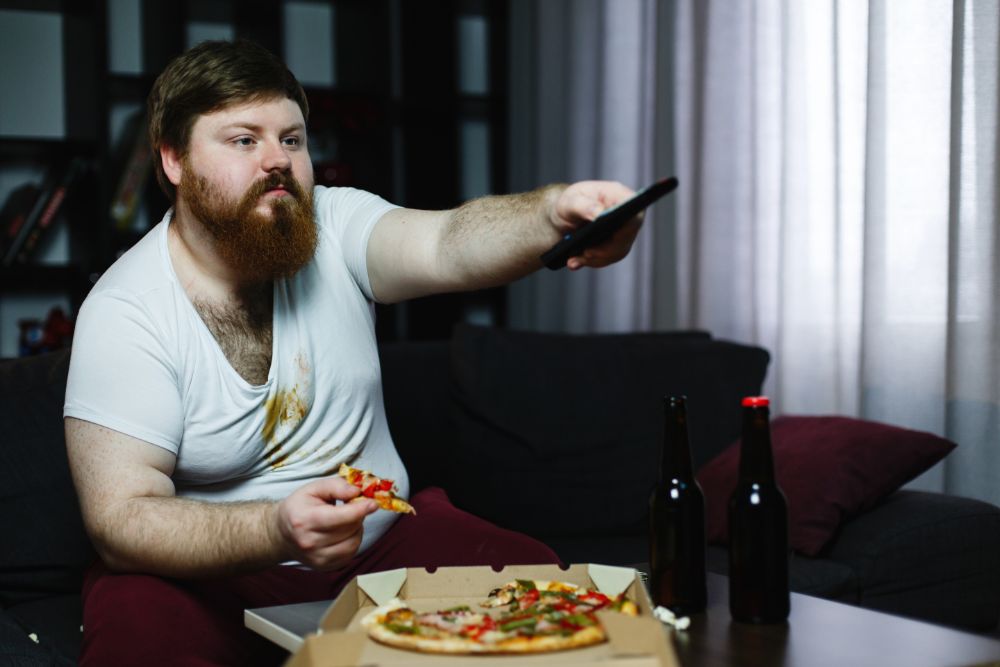 nezdravý životní styl, obézní muž sedí u televize a jí pizzu