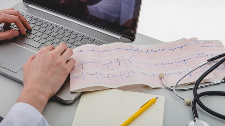 Lékař píše na notebooku a vedle leží záznam ekg a stetoskop